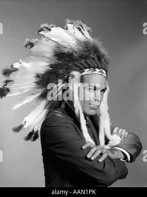 1960ER JAHRE MANN SÜFFISANTEN GESICHTSAUSDRUCK ARME VERSCHRÄNKT TRAGEN NATIVE AMERICAN INDIAN KOPFSCHMUCK Stockfoto