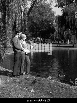 1960ER JAHREN PAAR MANN & FRAU FÜTTERN ENTEN AUF PARK TEICH UMGEBEN VON TRAUERWEIDE Stockfoto