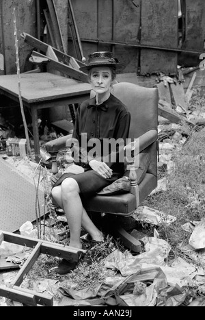 1970ER JAHRE ÄLTERE FRAU IM KLEID & HUT SITZT AUF LEDER DREHSTUHL UNTER TRASH & RUINEN MIT MÜRRISCHEN AUSDRUCK Stockfoto