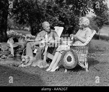 1930S 1940S IM RUHESTAND SENIOR PAAR SITZEN IM FREIEN RASEN STÜHLE MANN RAUCHEN PFEIFE FRAU STRICKEN MIT HUND Stockfoto