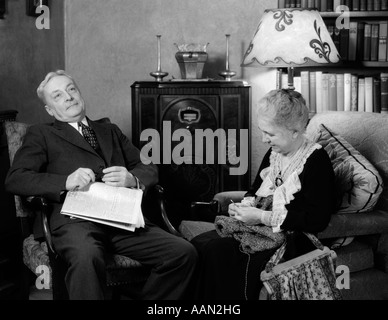 1930ER JAHRE ÄLTERES EHEPAAR SITZT IM WOHNZIMMER HÖREN RADIO FRAU STRICKEN MANN MIT ZEITUNG IN RUNDE Stockfoto