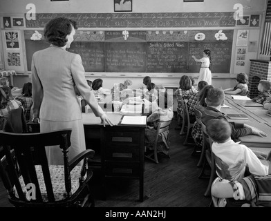 1950ER JAHRE MÄDCHEN AN DER VORDERSEITE DES KLASSENZIMMER MIT ZEIGER AUF DURCHLESEN SCHULNACHRICHTEN AUF TAFEL MIT LEHRER STEHEN AN DER REZEPTION Stockfoto