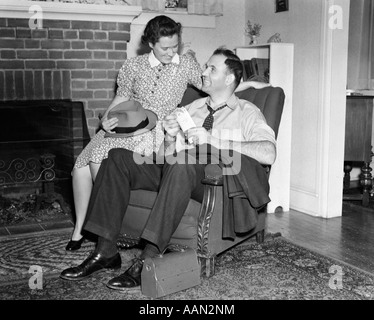 1940ER JAHREN LÄCHELND MANN SITZT IM STUHL PAY UMSCHLAG ZU HALTEN, WÄHREND FRAU AM ARM DER STUHL HÄLT SEINEN HUT MITTAGESSEN SITZT BOX ORIENTTEPPICH Stockfoto