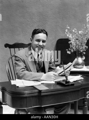 1930S 1940S PAPIERE GESCHÄFTSMANN AM SCHREIBTISCH BLICK AUF KAMERA HALTEN LÄCHELND DREITEILIGE ANZUG Stockfoto