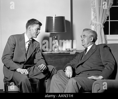 1950ER JAHRE JUNGER MANN SITZT IM WOHNZIMMER, IM GESPRÄCH MIT ÄLTEREN MANN Stockfoto