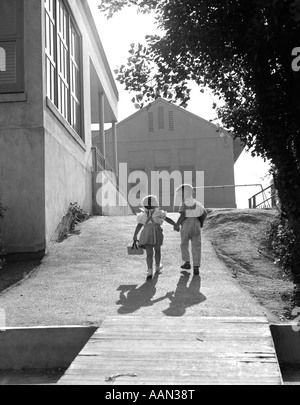 1940S 1950S ZWEI SCHULKINDER JUNGEN UND MÄDCHEN ZU FUß ZUR SCHULE HAND IN HAND Stockfoto