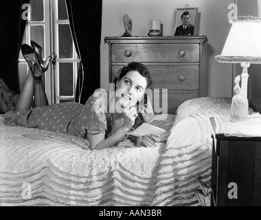 1940ER JAHREN BEKLEIDET VOLL, LÄCHELNDE FRAU AUF IHREM BETT MIT IHREN BEINEN, LIEST EINEN BRIEF IN IHREM SCHLAFZIMMER Stockfoto