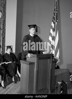 1950ER JAHREN VALEDICTORIAN GEBEN REDE AM PODIUM MIT ANDEREN ABSOLVENTEN SITZEN IM HINTERGRUND HÄLT DIPLOME Stockfoto