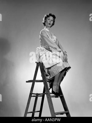 1950ER JAHREN FRAU IN SCHÜRZE MIT ÜBERTRIEBENEN AUSDRUCK SITTING ON TOP OF EINE TRITTLEITER, BLICK IN DIE KAMERA Stockfoto