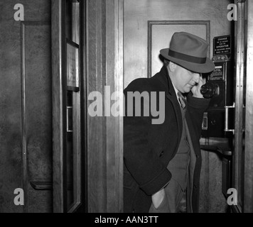 1930S 1940S GEKLEIDETE MANN IN FEDORA ANZUG UND MANTEL STEHT IN EINEM HOLZ GETÄFELTEN TELEFONZELLE Stockfoto