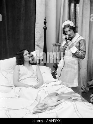 1920S 1930S ZWEI FRAUEN IM SCHLAFZIMMER EINE GEKLEIDET ALS MAGD AUF TELEFON ANDERE IM BETT LÄCHELTE ZOFE Stockfoto