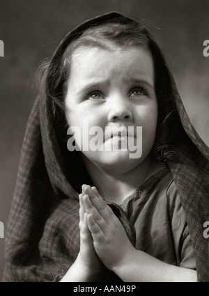 1930ER JAHREN ARME TRAURIGE KLEINE MÄDCHEN SCHAL ÜBER HAARE HÄNDE BETEND Stockfoto