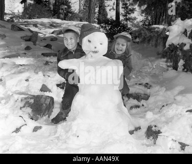 1950S 1960S LÄCHELNDEN JUNGEN UND MÄDCHEN GEBÄUDE EIN SCHNEEMANN ZUSAMMEN IM VERSCHNEITEN WALD Stockfoto