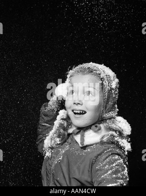 1950ER JAHREN KLEINE MÄDCHEN MIT KUNSTSCHNEE FLOCKEN FALLEN AM HUT UND HAAR WERFEN SCHNEEBALL IM STUDIO Stockfoto