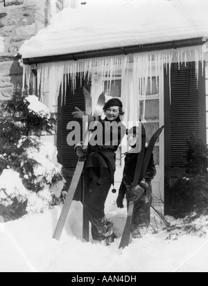 1940S 1930S MUTTER SOHN STEHEN VOR WINTERHAUS MIT EISZAPFEN HOLDING SKIER Stockfoto