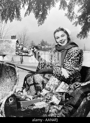 1950ER JAHREN LÄCHELNDE FRAU BLICK IN DIE KAMERA REITEN IN SCHLITTEN TRAGEN LEOPARD HAUT PELZMANTEL IN MAILBOX MIT PAKETEN Stockfoto