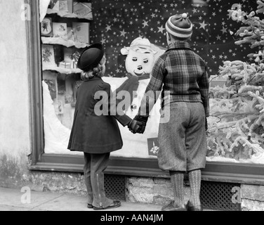 1930ER JAHREN WIEDER DEKORIERT BLICK JUNGEN UND MÄDCHEN HAND IN HAND AUF DER SUCHE AT WEIHNACHTS-FENSTER Stockfoto