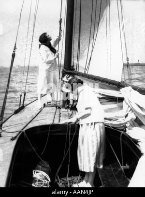 1920ER JAHREN ZWEI FRAUEN AUF DEM DECK DES CATBOAT IM COCKPIT ZIEHEN SEILE ANDERE STEHEND DURCH MAIN MAST ZIEHEN SEIL RAISE MAIN SAIL Stockfoto