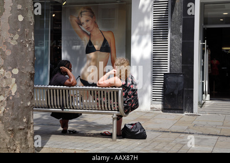 zwei Frauen sitzen auf einer Bank in Croydon, England mit einem Poster von Kate Moss im Hintergrund Stockfoto