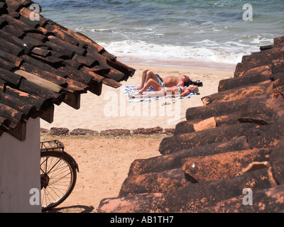 Paar Sonnen Sie sich am Rand des Wassers am Strand eingerahmt von Ziegeldächern Anjuna Goa Indien Stockfoto