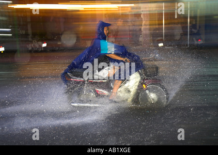 Motorradfahrer in Kap wirft Spray Reiten entlang überflutet De Tham Street Pham Ngu Lao Bezirk Ho Chi Minh City, Vietnam Stockfoto