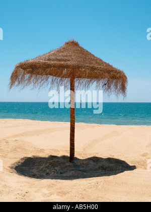 THATCHED Sonnenschirm wirft einen Schatten um die Mittagszeit auf einem leeren Sandstrand mit blauen Meer Himmel und Horizont. Algarve Portugal Europa Stockfoto