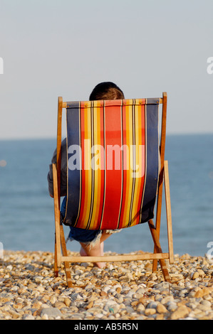 RÜCKANSICHT DER MANN SITZT IM LIEGESTUHL AM KIESSTRAND MIT BLICK AUF DAS MEER Stockfoto