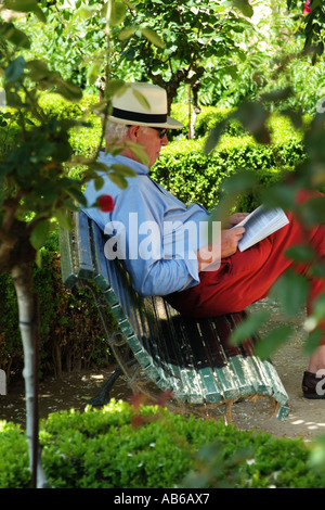 Mann im Strohhut und Freizeitkleidung einen Reiseführer lesen. Sitzen auf einer Gartenbank. Europäische Stockfoto