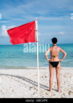 Frau stehend auf Te Strand in der Nähe eine rote Fahne auf dem Meer, was bedeutet, dass Schwimmen ist gefährlich und verboten Stockfoto