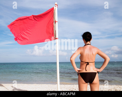 Frau stehend auf Te Strand in der Nähe eine rote Fahne auf dem Meer, was bedeutet, dass Schwimmen ist gefährlich und verboten Stockfoto