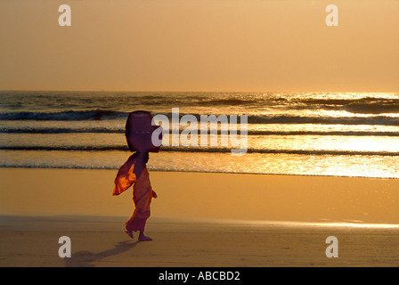 Goan Frau mit Korb auf Kopf Spaziergänge am Sandstrand Benaulim. Sonnenuntergang Reflektionen im Meer und Brandung, Salcete Goa Indien Stockfoto