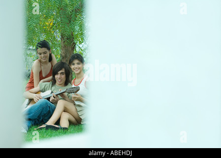 Gruppe junger Freunde von Baum sitzen, während man Gitarre spielt Stockfoto