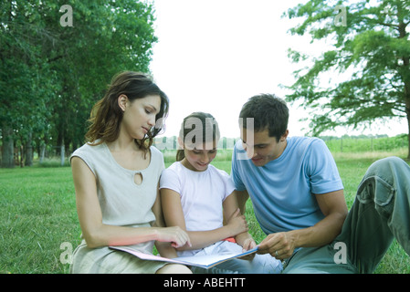 Mädchen lesen Buch mit Eltern, sitzen auf dem Rasen Stockfoto