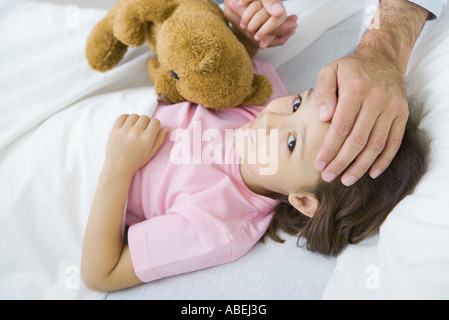 Mädchen liegend im Bett mit Teddybär, Mann hielt ihre Hand über die Umsetzung von anderer Hand auf der Stirn Stockfoto