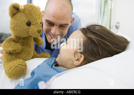 Mädchen im Krankenhausbett liegend, Krankenschwester, Teddybär hochhalten Stockfoto