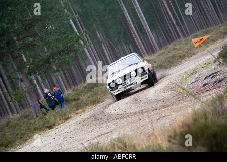 Weißen Ford Escort Rallye-Auto fahren Action Spritzen Kies Steinen Dreck und Schlamm, wie sich herausstellt eine Ecke Kiefern sehen 2 Personen Stockfoto