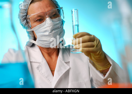 Weibliche Laborantin mit Schutzbrille Handschuhe Maske und Haarnetz Überprüfung Flüssigkeitsvolumen im Messzylinder auf blauem Hintergrund Stockfoto