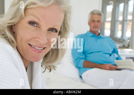 Paar im Schlafzimmer, Frau sucht Mann mit einem Buch im Hintergrund in die Kamera, Stockfoto