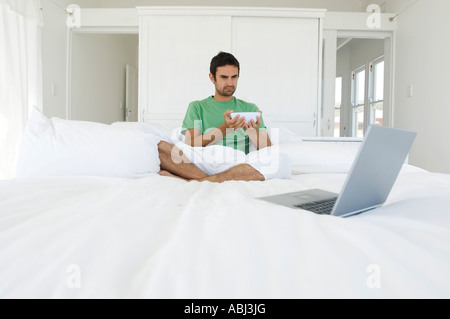 Junger Mann sitzt mit gekreuzten Beinen auf einem Bett, Blick auf laptop Stockfoto