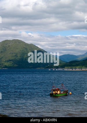 Ein kleine grüne und gelbe Fischerboot vor Anker im Loch Crerans mit einem Jachthafen und Hügeln hinter Highlands Scotland UK Stockfoto