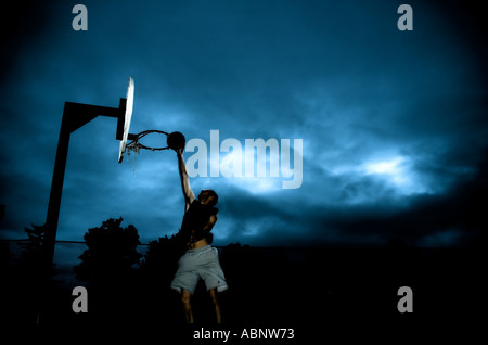 Eine Silhouette eines jungen Mannes einen Basketball dunking. Dunkler Himmel mit Licht spähen durch im Hintergrund. Stockfoto