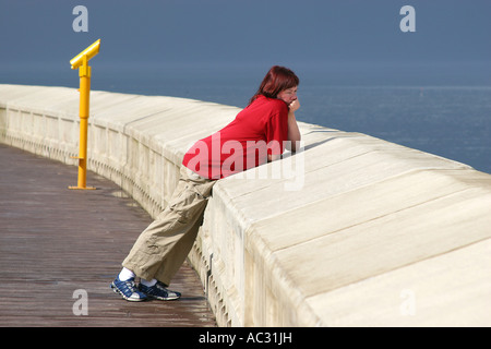 Schwangere junge Frau lehnt sich am Deich mit Blick auf das Meer mit gelben Teleskop im Hintergrund. Stockfoto