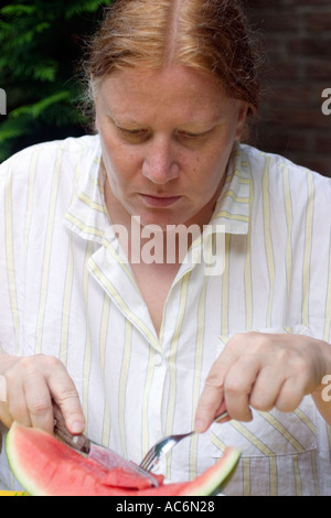 Mitte gealterte Frau lässig gekleidet, Wassermelone mit Messer und Gabel zu essen und ein Buch zu lesen, im Sommer im Garten Stockfoto