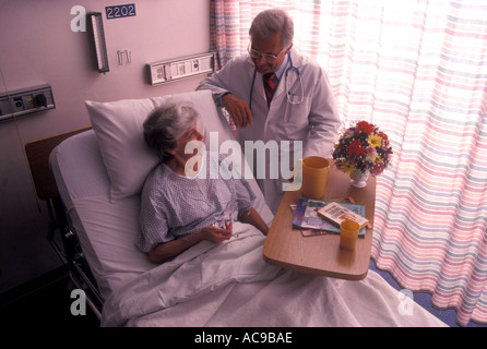 Einen männlichen Arzt mit weißen Lab Mantel Stethoskop Brille stehend am Bett einer Patientin in ein Krankenhaus Zimmer Deko Stockfoto