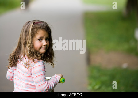 Kleines Mädchen im Park auf dem Roller hinter in die Kamera schauen Stockfoto