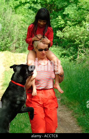 Herr PR schwarzen Hund und junge dunkelhaarige Mädchen im Alter von 6 Jahren in der Natur sitzen auf Schultern von einem mittleren Alter Frau Stockfoto