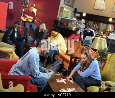 Junge Erwachsene in einer Kaffeebar herumsitzen und reden, spielen Domino oder Dame oder auf einem Laptop arbeiten, wie ein Gitarrist auf der Bühne spielt Stockfoto