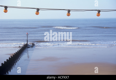 Eine Reihe von orange Glühbirnen über den Himmel über ein ruhiges blau graue Meer mit plätschernden Wellen und hölzernen Buhne aufgereiht Stockfoto