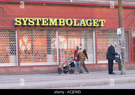 Die Vorderseite eines den Systembolaget-Läden, das Retail-Monopol für den Verkauf von Alkohol Wein Bier und Spirituosen, eine gelbe Leuchtreklame und des Eisernen Draht geschlossen Soder im Einkaufszentrum Ringen. Ein Mann mit einer Plastiktüte auf der Suche nach etwas Sinnvolles in die Mülltonne. Ein junges Paar mit einem Kind in einem Kinderwagen trolley Perambulator Kinderwagen Buggy Kinderwagen vorbeigehen im Shop Fenster Stockholm, Schweden, Sverige, Europa suchen Stockfoto