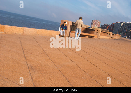 Eine Frau sitzen trinken Mate Kräutertee und ein Mann zu Fuß auf dem Bürgersteig, auf dem Spaziergang am Flussufer am Meer entlang des Flusses Rio De La Plata Ramblas Sur, Gran Bretagna und Republica Argentina Montevideo, Uruguay, Südamerika Stockfoto